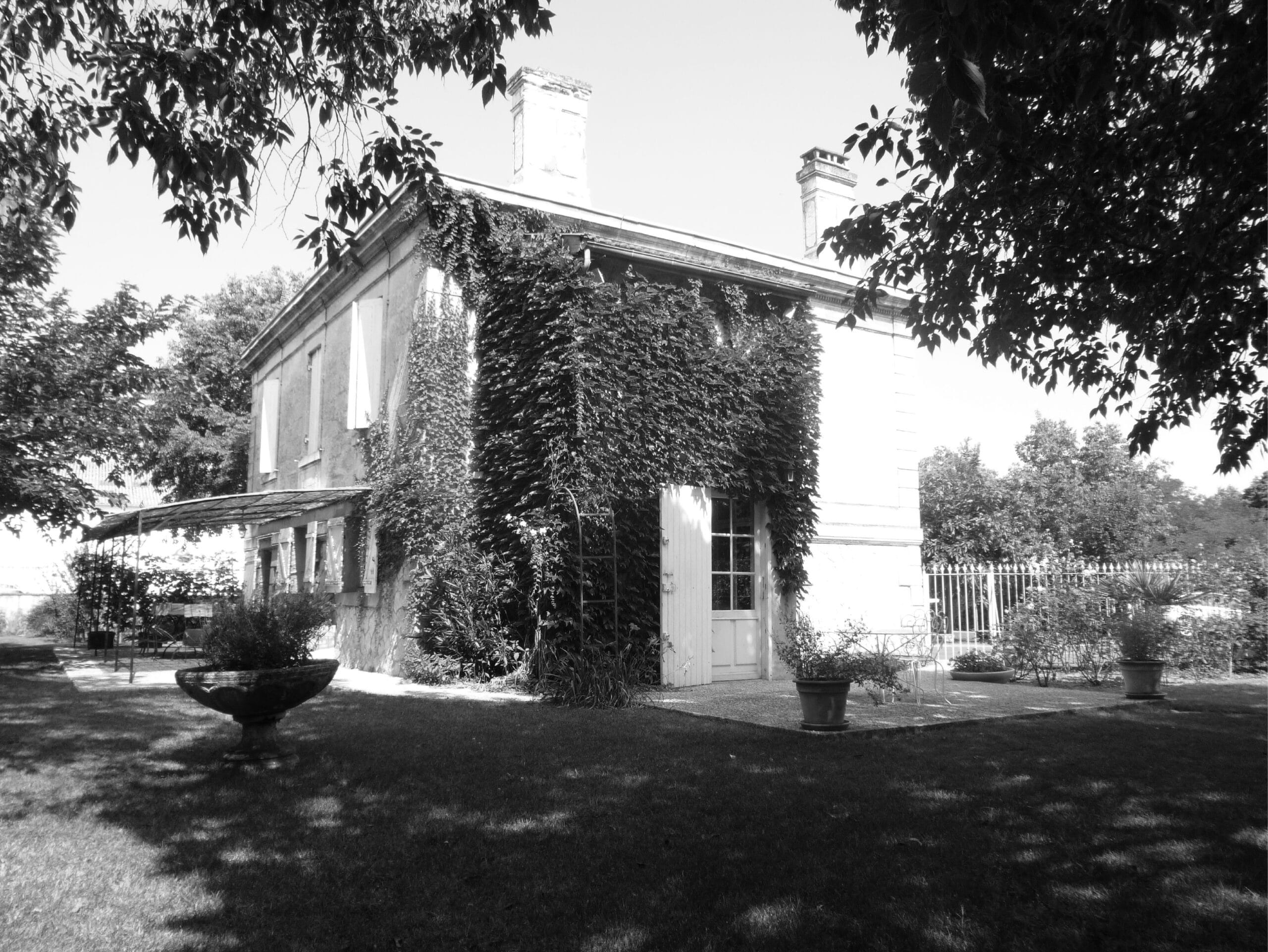 Façade en noir et blanc d'une maison de maître élégante, couverte de lierre. Le jardin d'été ombragé présente une pelouse et de grandes vasques.