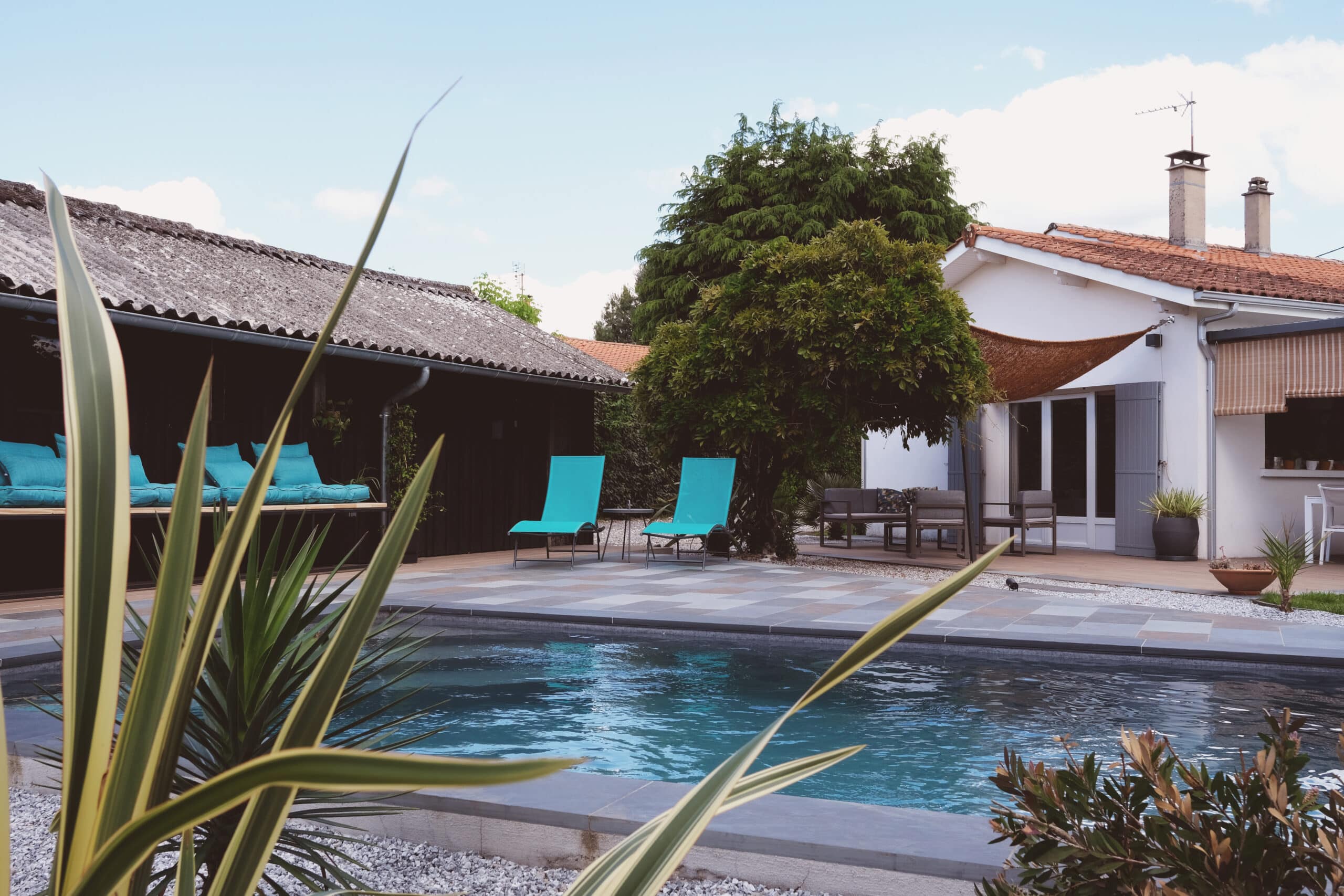 Piscine moderne et jardin aménagé pour détente estivale Piscine moderne avec chaises longues bleu turquoise et mobilier extérieur, devant une maison blanche et une dépendance au toit ondulé.