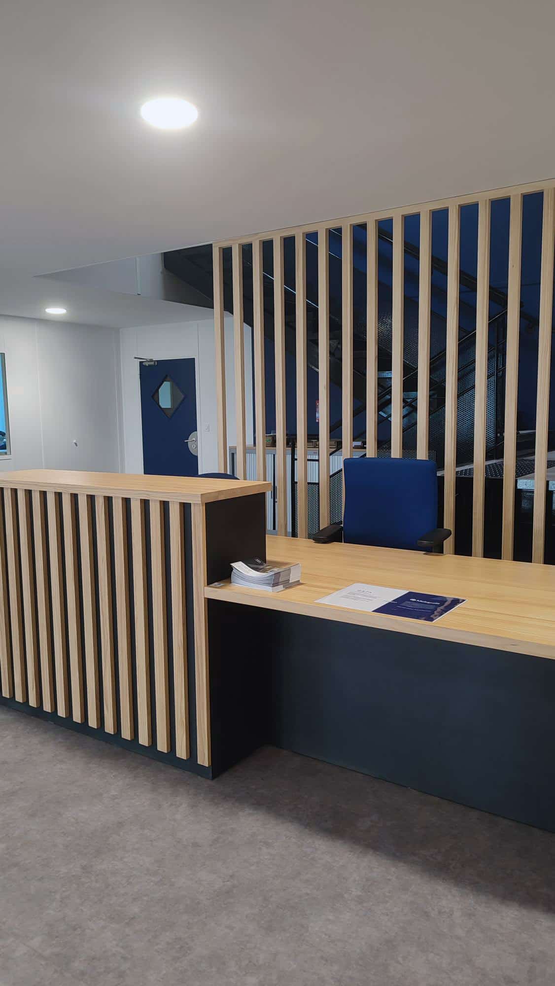 Accueil moderne avec comptoir bois clair à lattes verticales, base bleu nuit, et séparateur en claustra bois devant un escalier sombre. Porte bleue visible.
