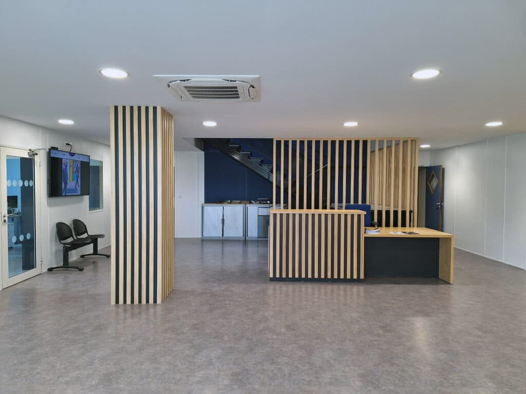 Hall d'accueil moderne avec comptoir en lattes de bois clair, écran affichant 14:50, et escalier bleu visible derrière la structure.