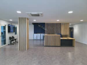 Hall d'accueil moderne avec comptoir en lattes de bois clair, écran affichant 14:50, et escalier bleu visible derrière la structure.