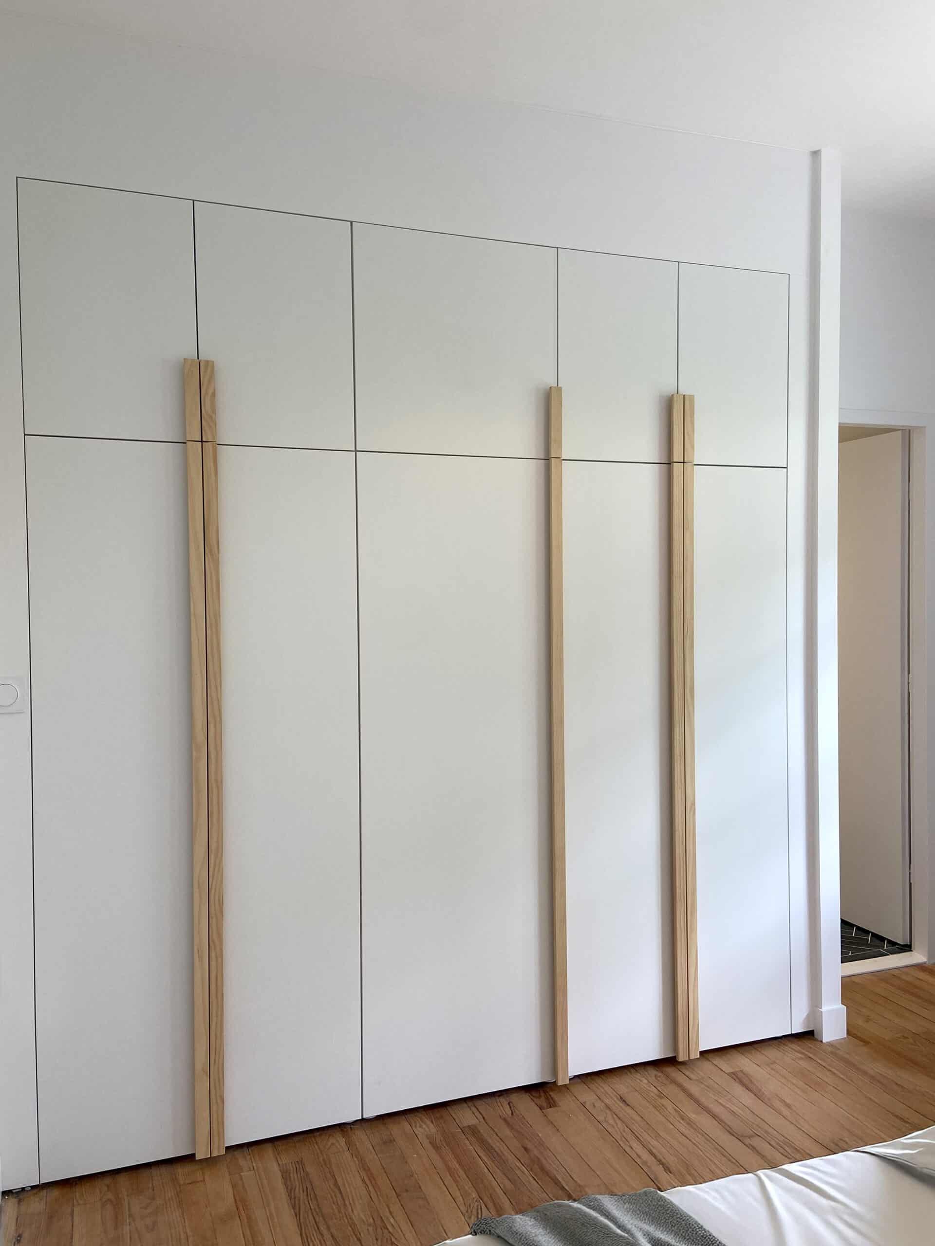 Garde-robe moderne blanche à quatre portes, avec poignées verticales en bois clair, installée près d'un parquet.