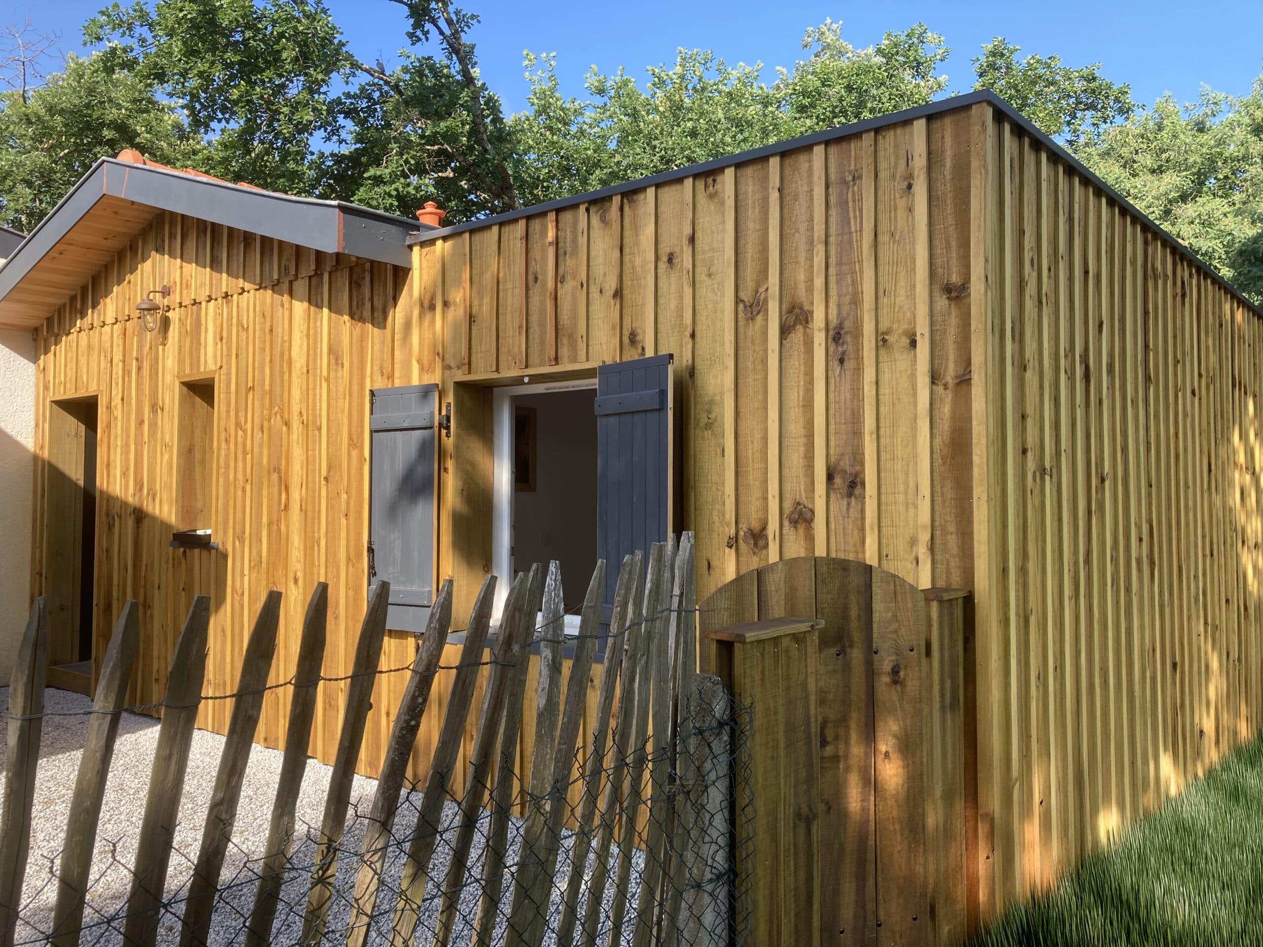 Cabane en bois brun clair avec bardage vertical, volets gris ouverts, devant une clôture rustique et des arbres verts.