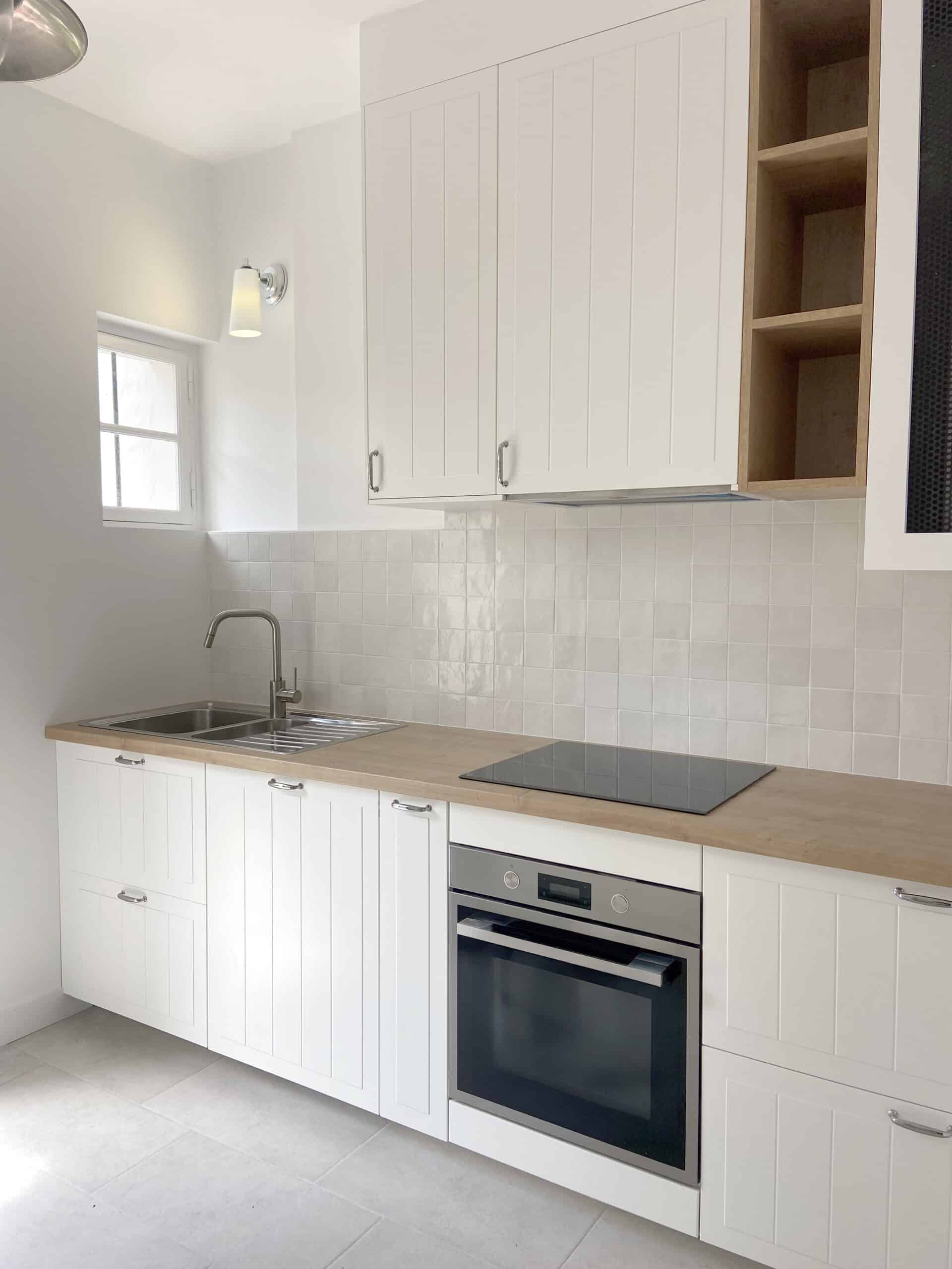 Cuisine moderne et lumineuse avec armoires blanches shaker, plan de travail en bois clair, carrelage mural blanc brillant et four encastré.