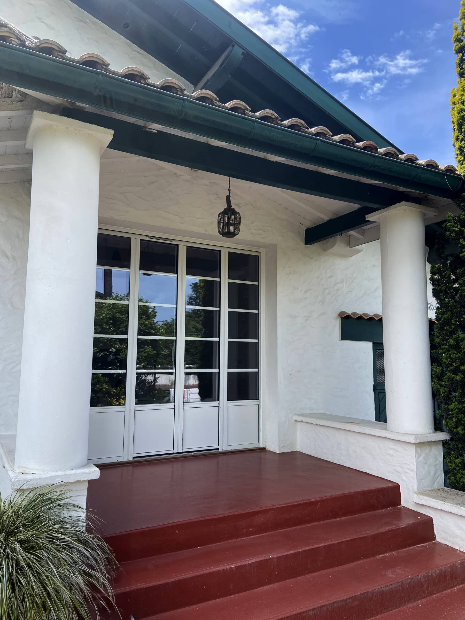 Entrée d'une maison blanchie à la chaux avec une terrasse rouge, un porche soutenu par des colonnes blanches et des portes vitrées à meneaux.