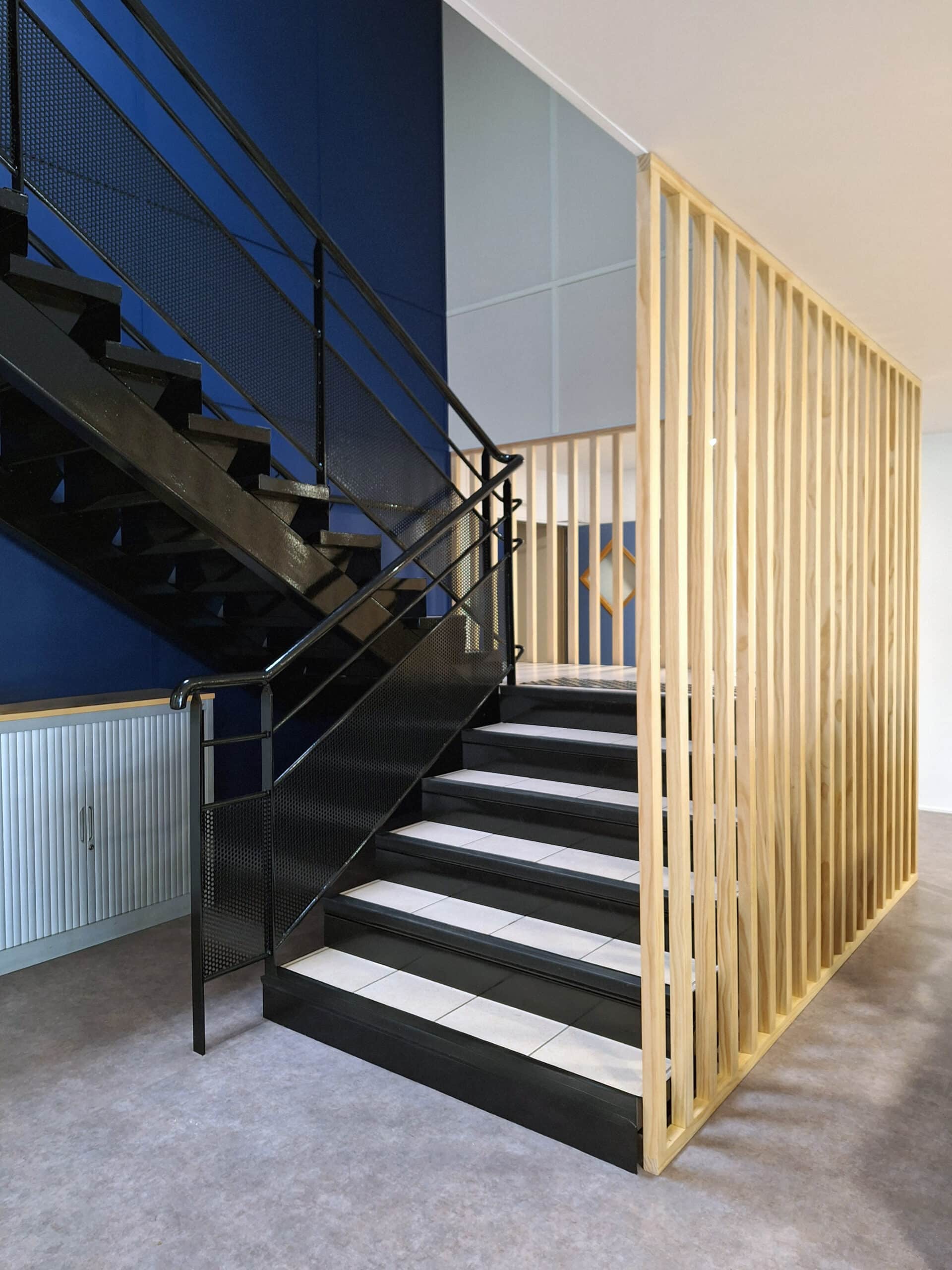 Escalier moderne noir et blanc à côté d'un claustra en bois clair, avec un mur bleu foncé et un meuble gris à stores.