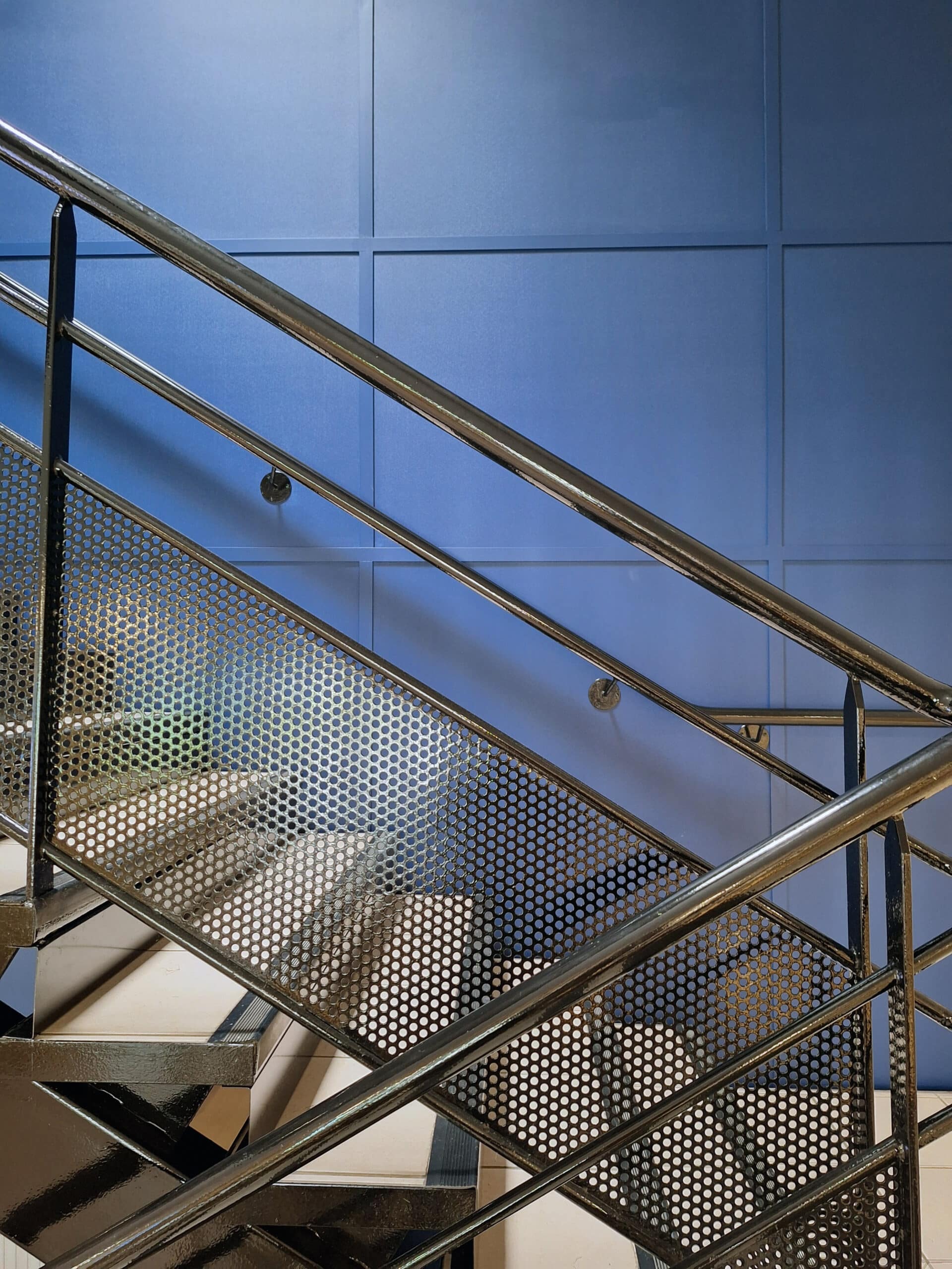 Escalier moderne avec main courante métallique brillante et garde-corps en tôle perforée, contrastant avec un mur bleu à panneaux.
