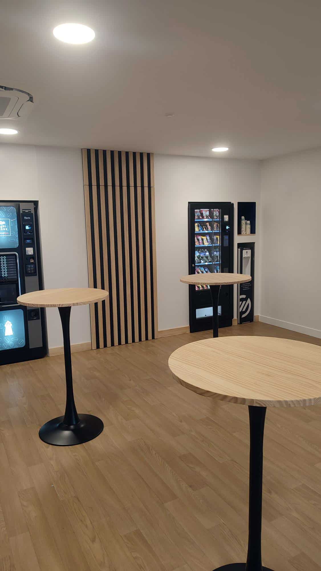 Espace détente moderne avec trois tables hautes à plateaux bois clair et pieds noirs, entourant des distributeurs automatiques Necta et un distributeur d'eau.
