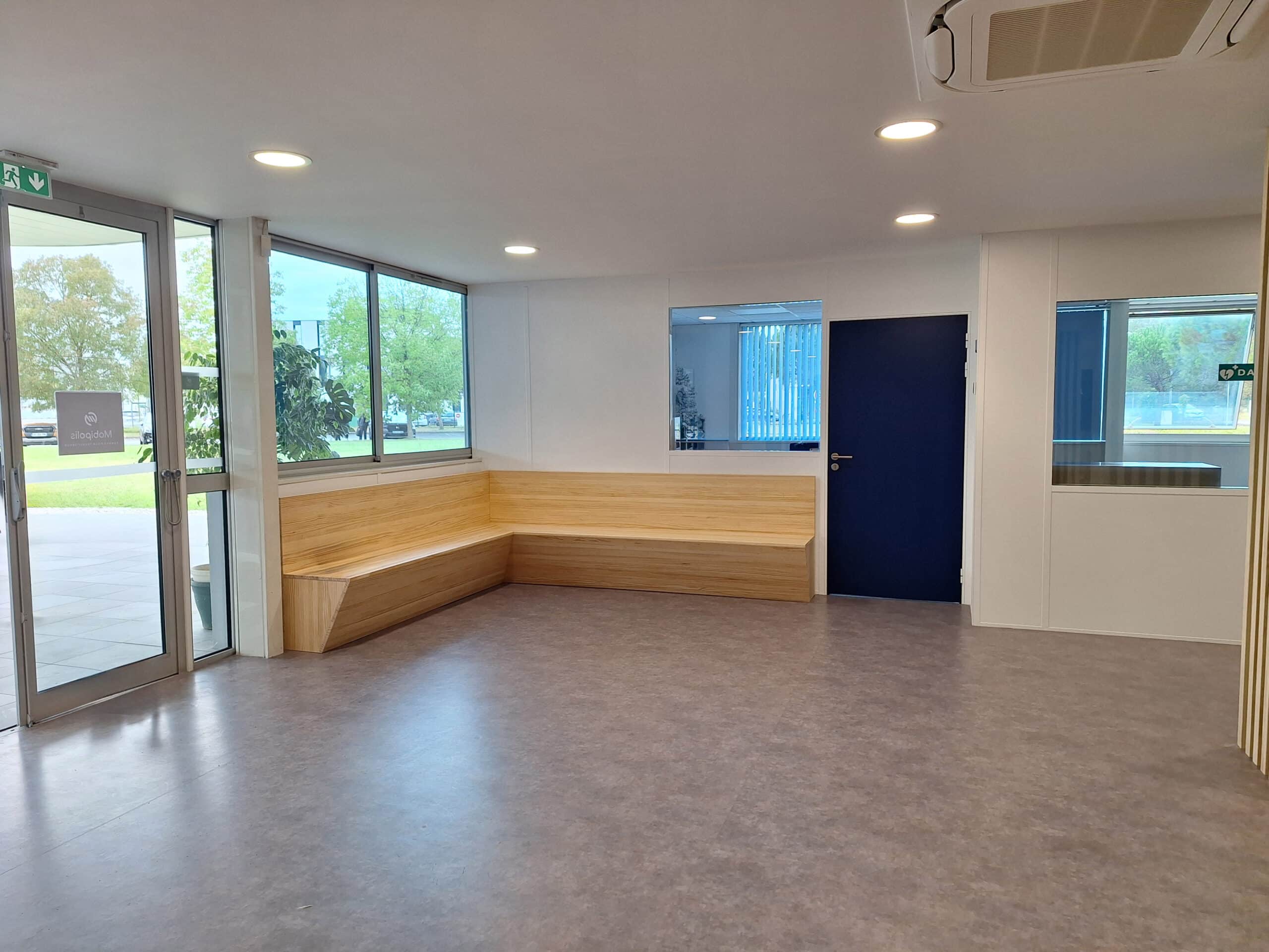 Hall d'accueil moderne avec banquette d'angle en bois clair, porte de sortie de secours (icône verte) et porte pleine bleu marine.