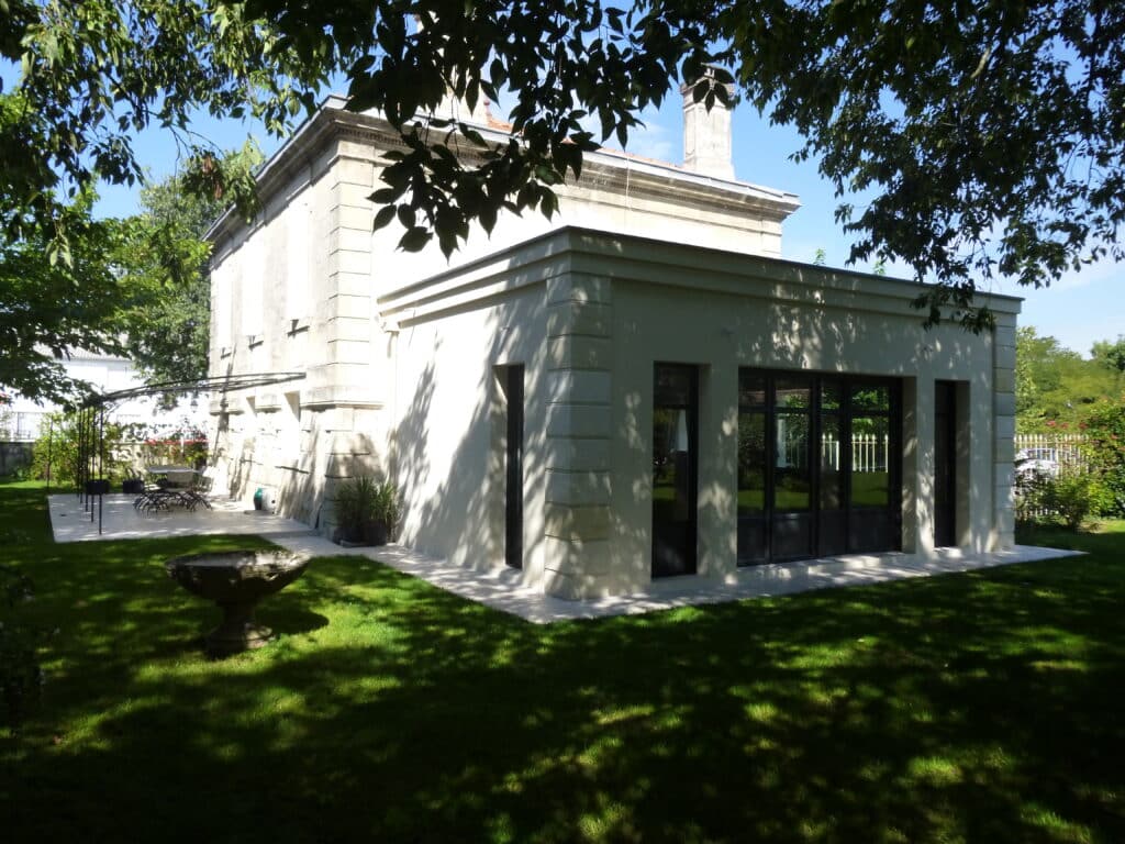 Belle extension de maison ancienne en pierre avec grandes baies vitrées noires, vue depuis un jardin verdoyant et ombragé par des arbres.