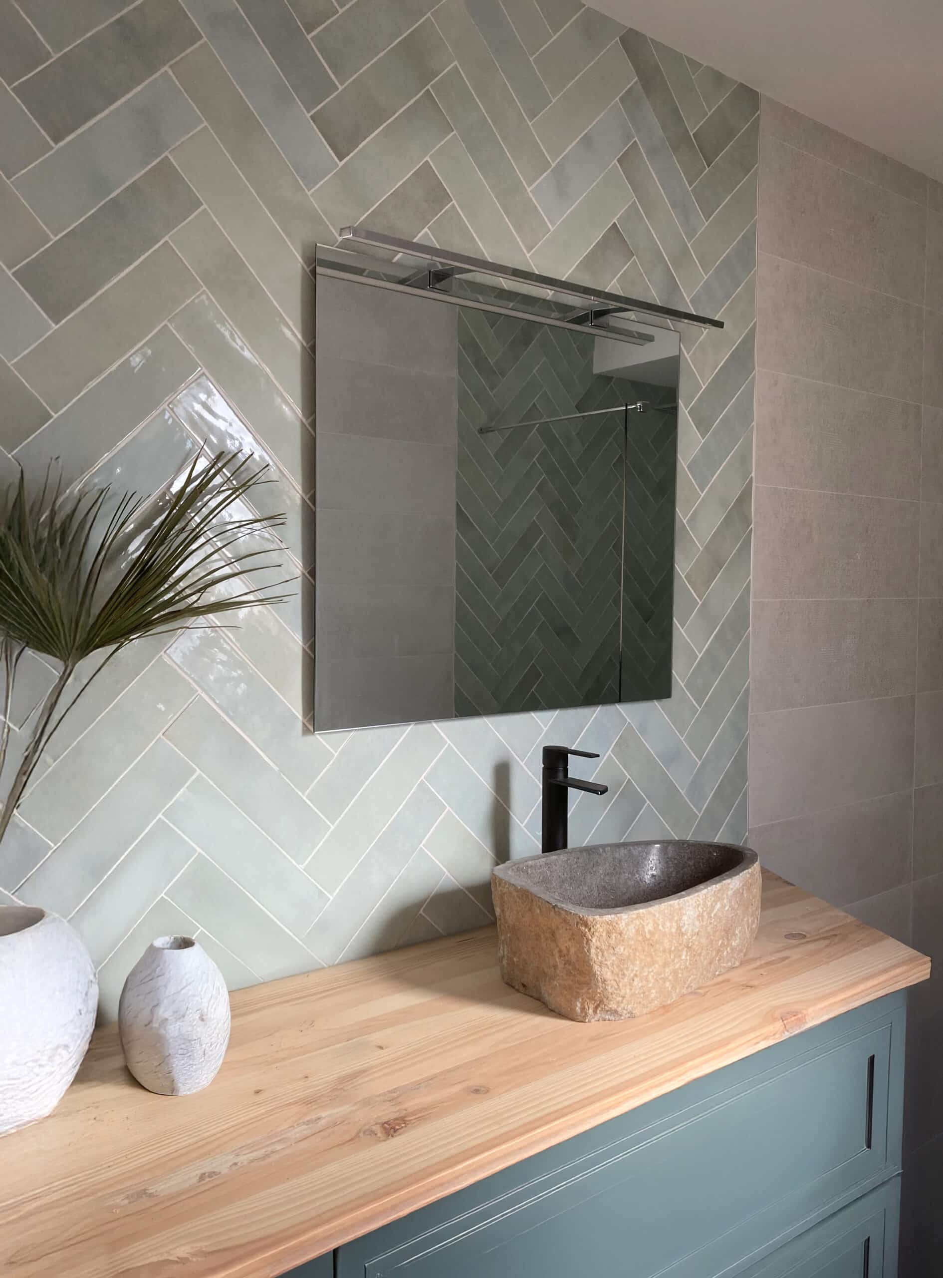 Salle de bain moderne avec carrelage mural vert sauge à motif chevron, meuble-lavabo en bois clair et vasque en pierre brute.