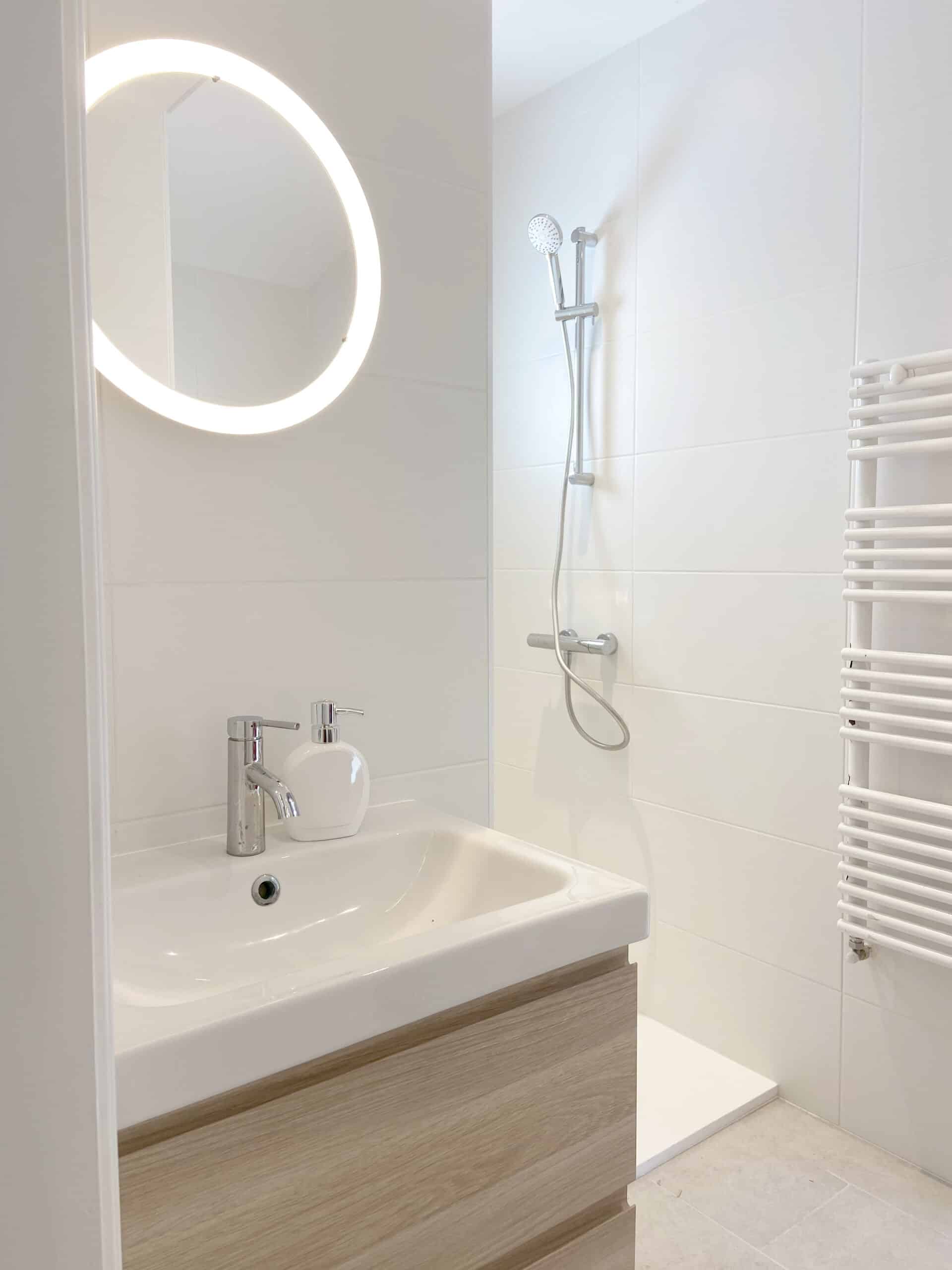 Salle de bain moderne et lumineuse avec vasque en bois clair, miroir rond rétroéclairé, douche à l'italienne et sèche-serviettes blanc.