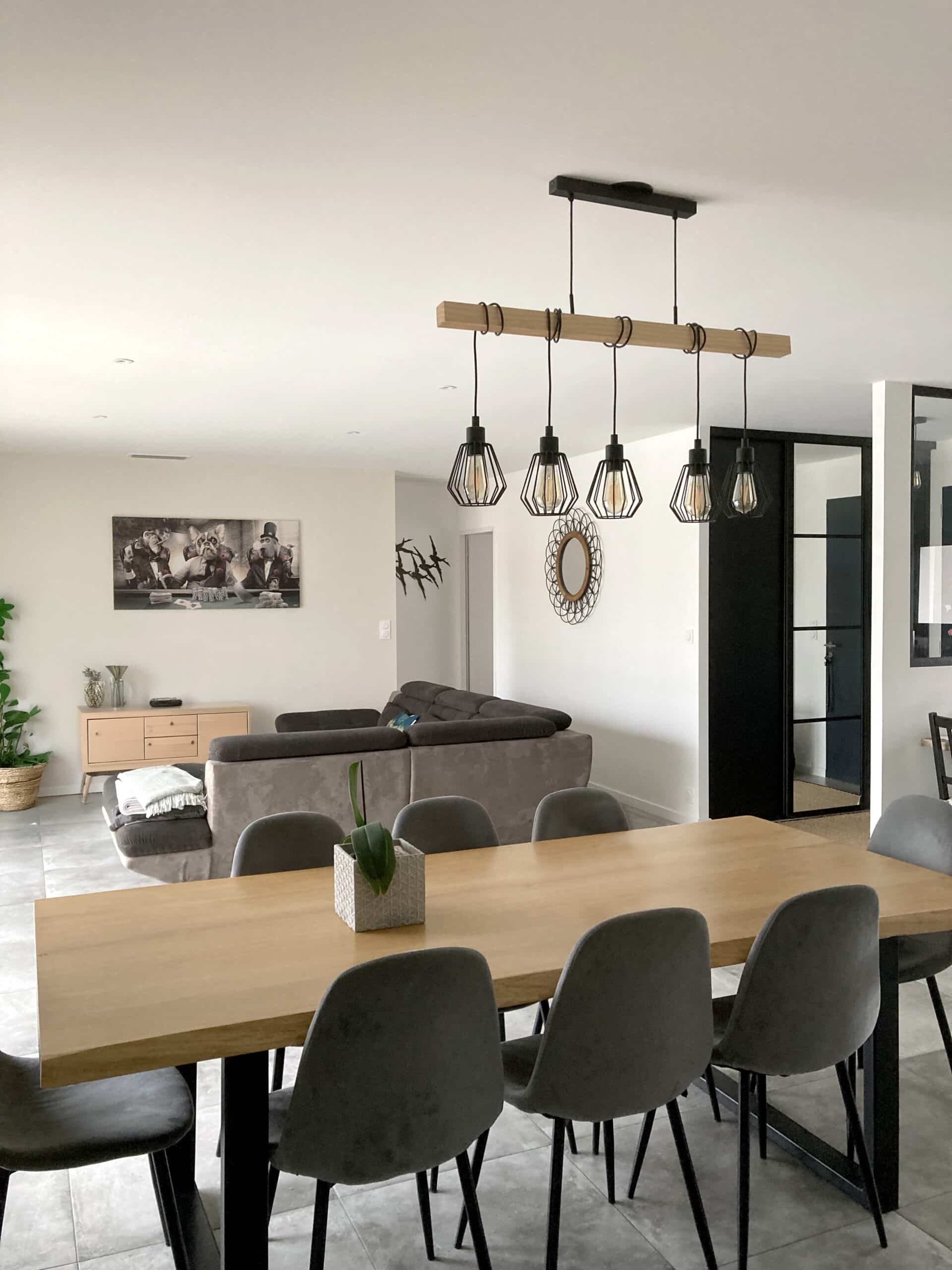 Salle à manger moderne avec table en bois clair, chaises grises, lustre industriel à poutre bois et aperçu d'un salon avec canapé et tableau d'animaux anthropomorphes.