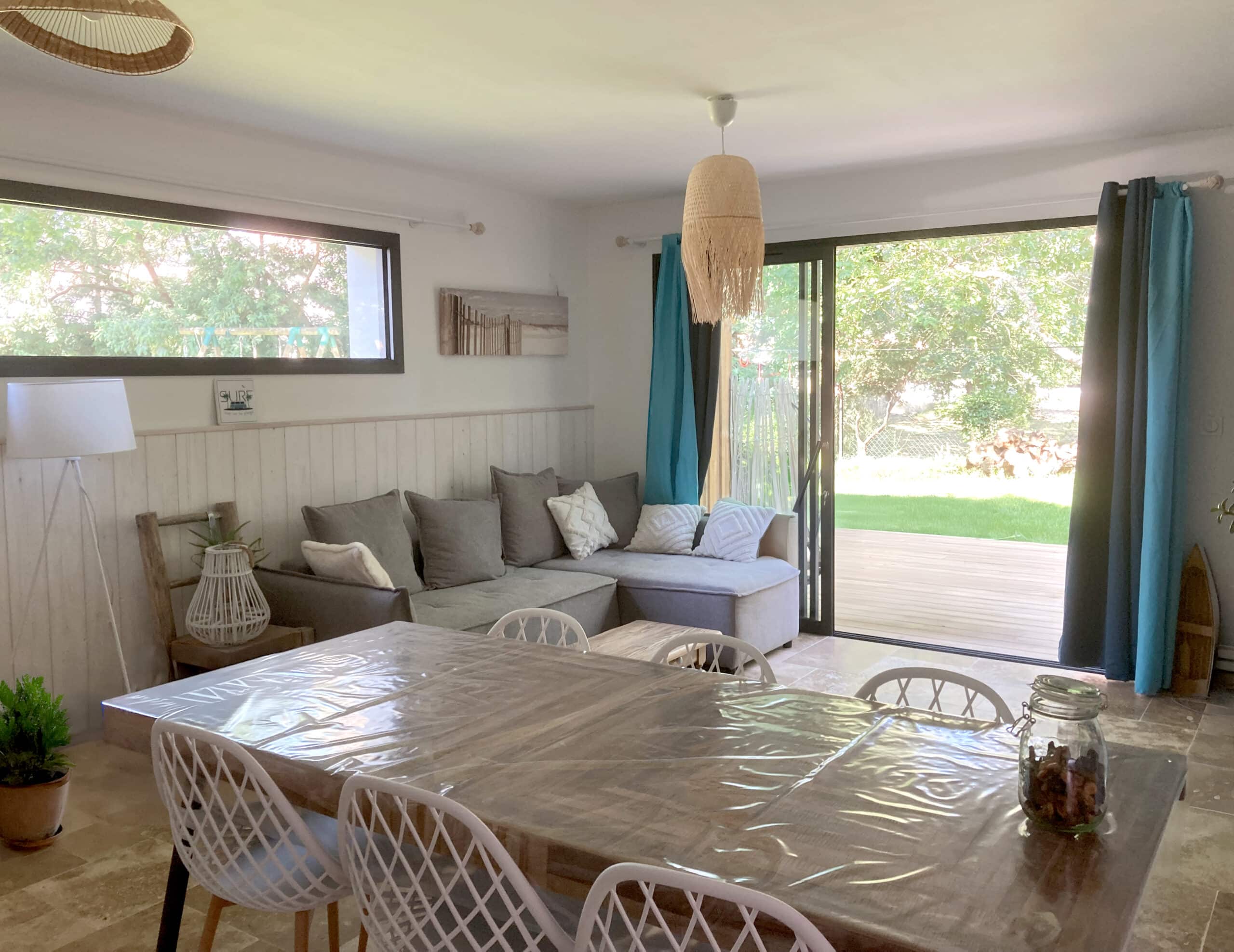 Salle à manger/salon lumineuse avec table recouverte de plastique, canapé gris, chaises blanches et vue sur terrasse et jardin verdoyant. Tableau « SURF » visible.