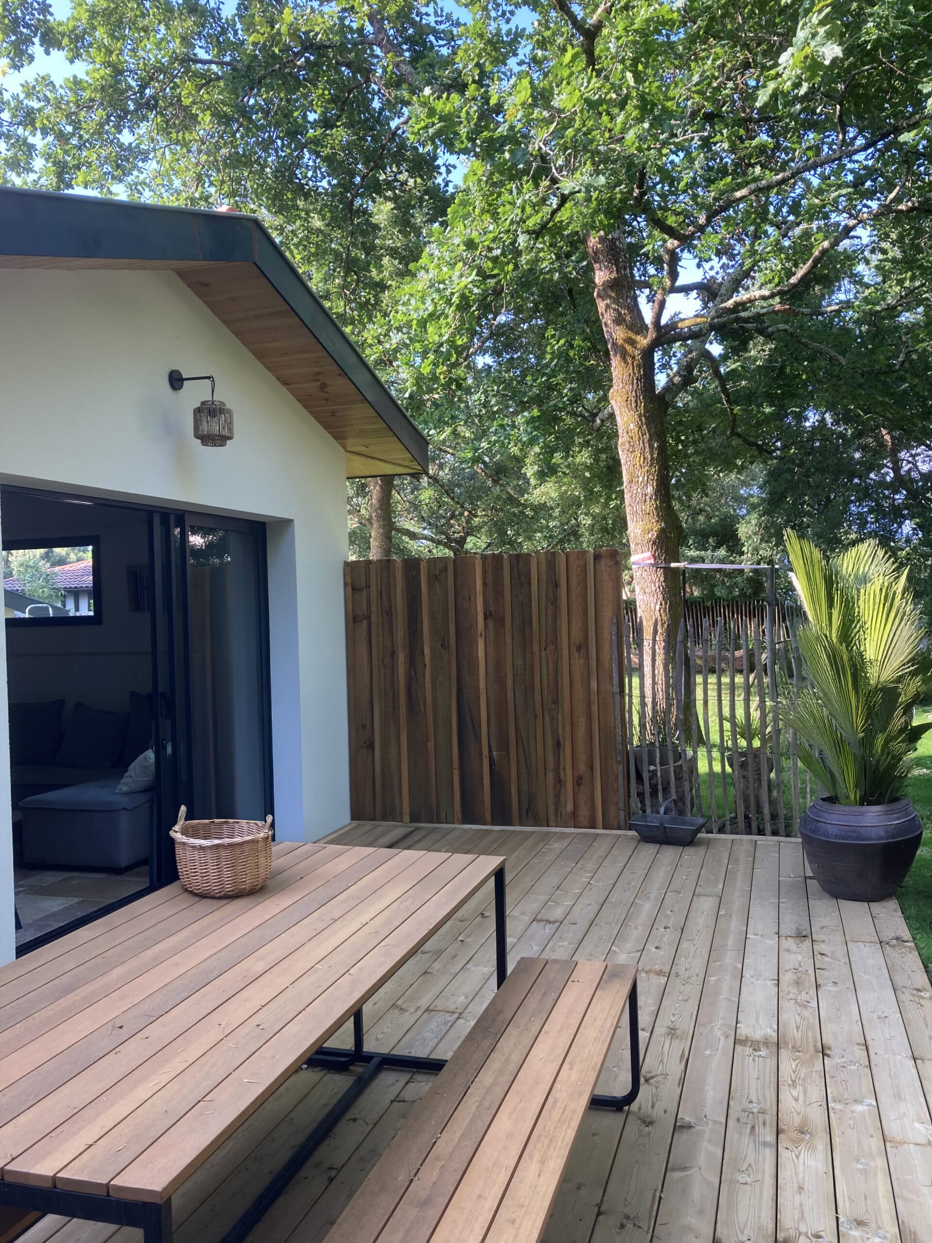 Terrasse en bois moderne devant une maison claire, avec une table et bancs en bois, éclairée par le soleil et entourée de verdure et d'arbres.