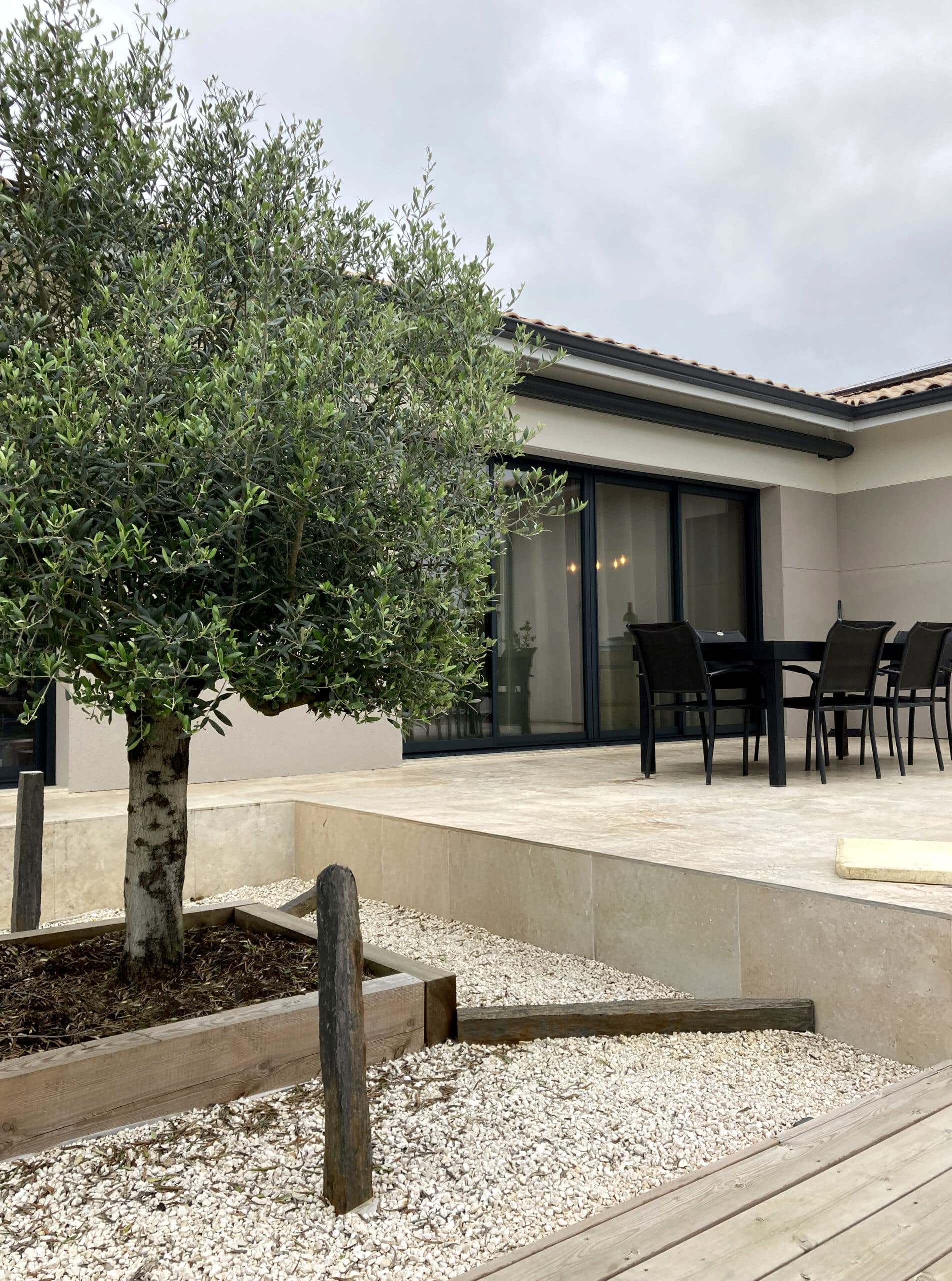 Olivier en pot à côté d'une terrasse carrelée avec salon de jardin noir, sous un ciel gris et nuageux.