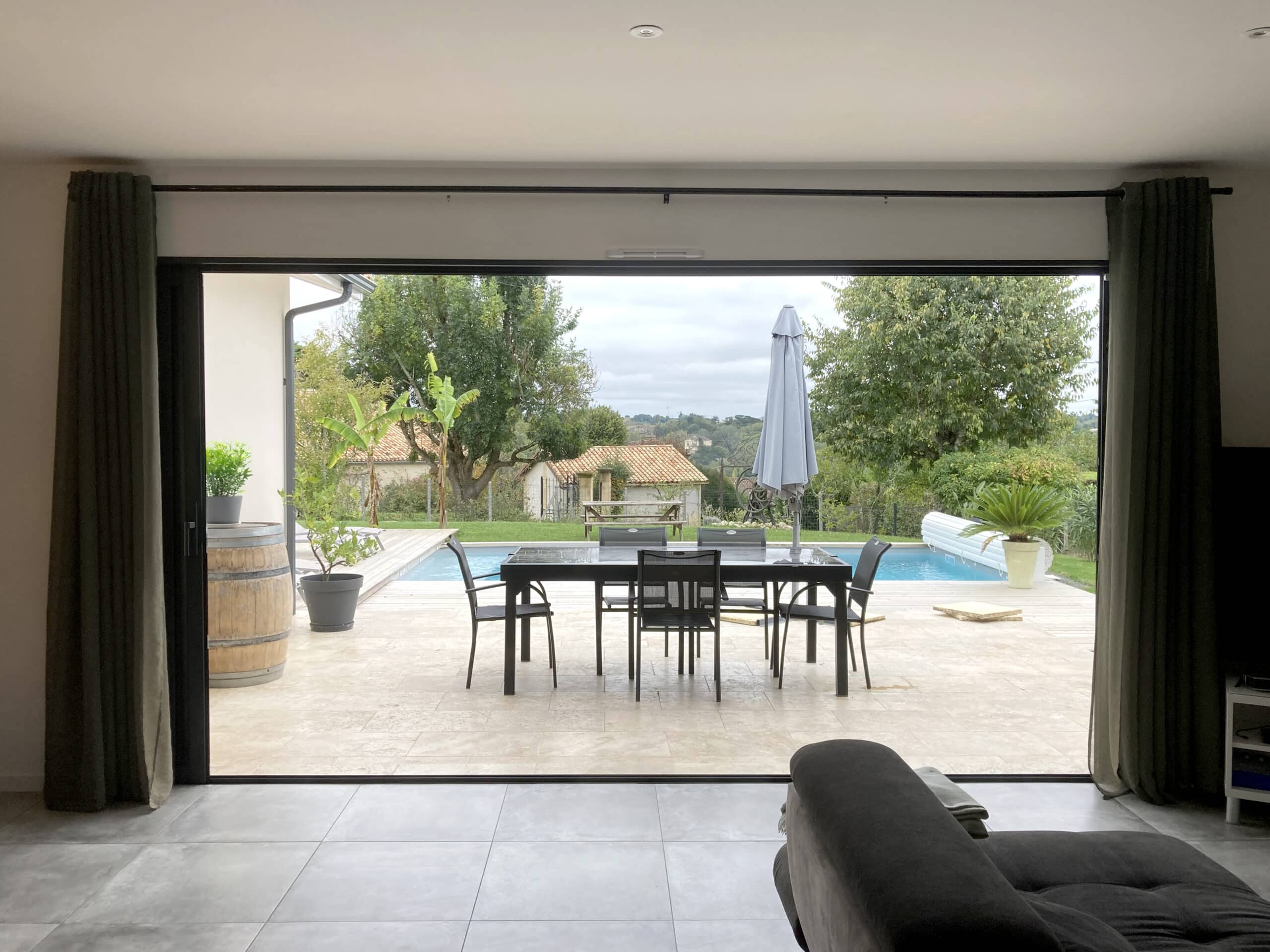 Vue d'un salon moderne vers l'extérieur: grandes baies vitrées ouvertes sur une terrasse avec mobilier de jardin, piscine et verdure.